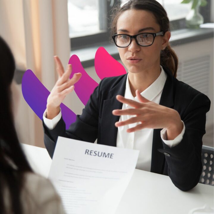 A professional woman wearing glasses and a business suit is seated at a desk during a law firm interview. She is gesturing with her hands as she speaks, while the interviewer, whose back is visible, holds a resume. The background suggests a modern office environment with natural lighting.