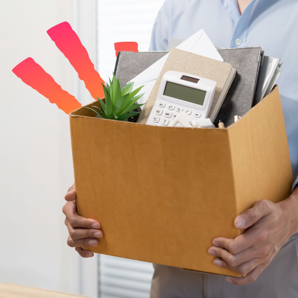 Person holding a cardboard box filled with personal belongings, including a plant, calculator, and documents, symbolizing being laid off.