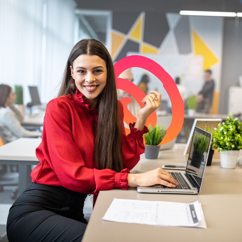 Smiling professional woman in a red blouse working on a laptop in a modern office, showcasing confidence and positivity in the workplace.