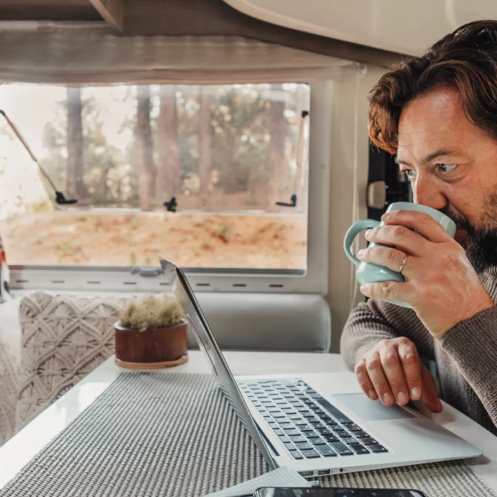 Man working on laptop computer inside a camper van with nature view outside the window.