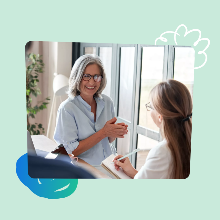 Seasoned Female professional mentoring a female apprentice; apprentice is listening while taking notes inside an office.