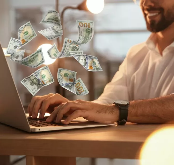 Closeup view of man using laptop at table and flying dollars making money