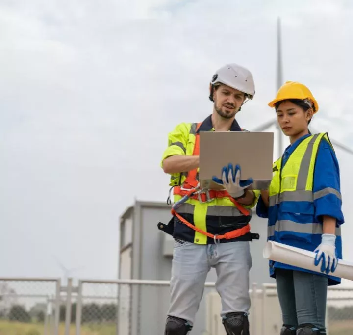 Male and female public utilities engineer in uniform with helmet safety using laptop discussing inspection and maintenance of wind turbine in wind farm to generate electrical energy.