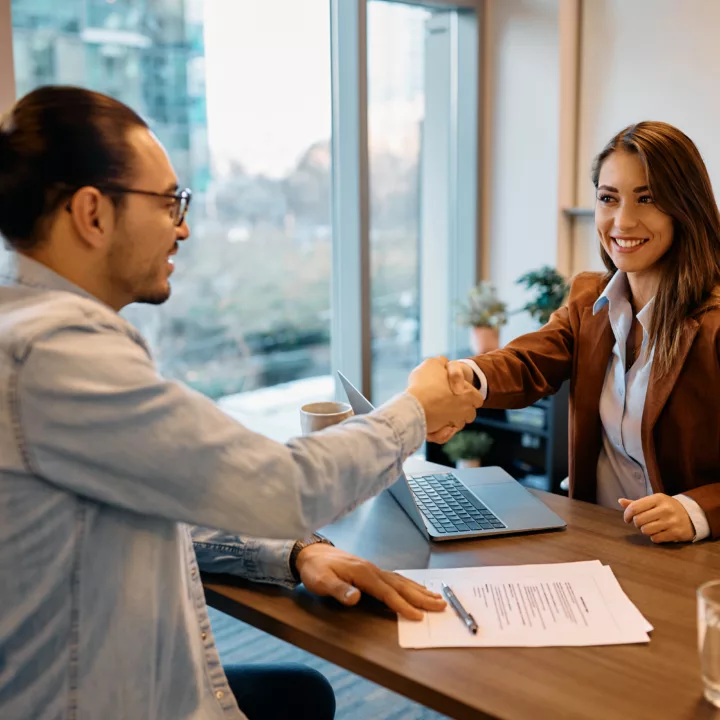 Female hiring manager smiling shaking male candidate's hand during an interview sitting at a desk in an office with resume laying out. Candidate is showing great interview body language by sitting up tall and giving a firm handshake.