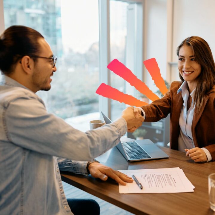 Female hiring manager smiling shaking male candidate's hand during an interview sitting at a desk in an office with resume laying out. Candidate is showing great interview body language by sitting up tall and giving a firm handshake.