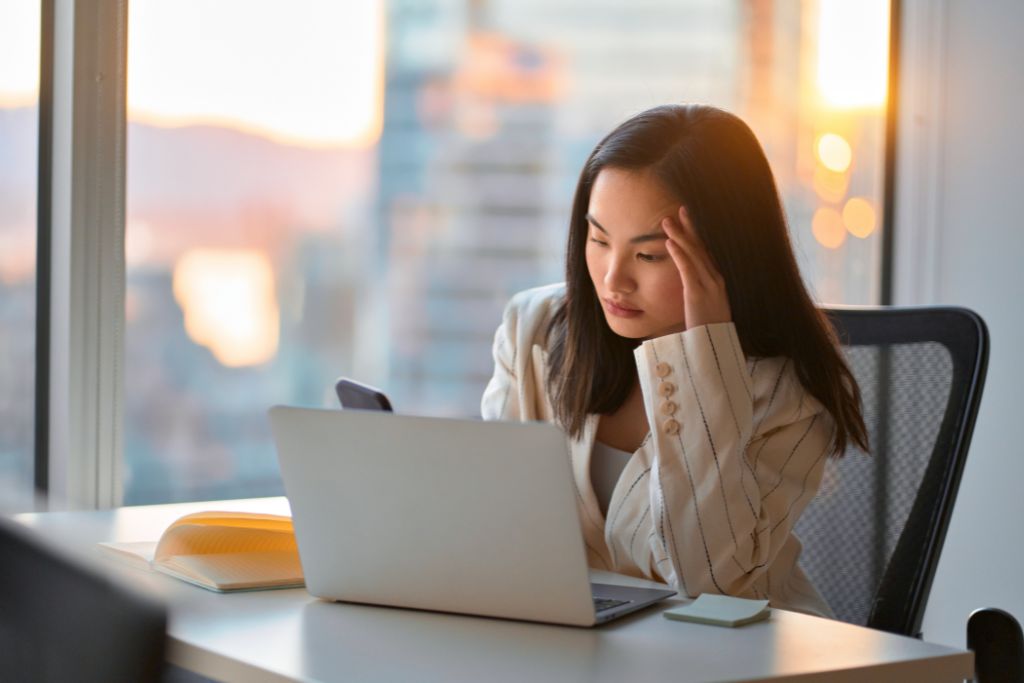 Woman professional looking unhappy sitting at her desk in an office resting her head on her hand looking at her phone behind a laptop wondering if her career plateaued.