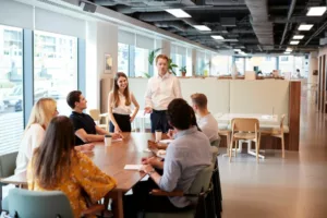 Professional working on his communication skills at business meeting. Professional standing up with a female coworker and six other coworkers sitting around a table