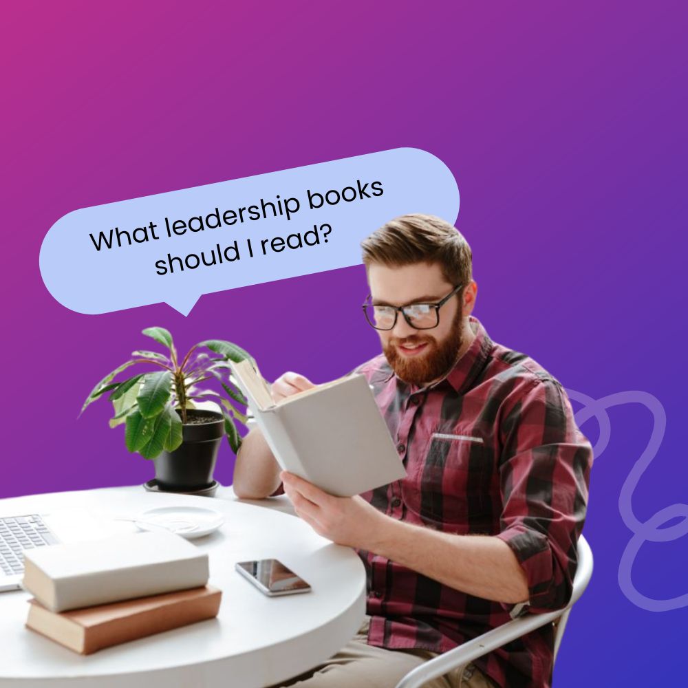 Male leader in a flannel reading a leadership book at his desk next to a laptop and pile of books