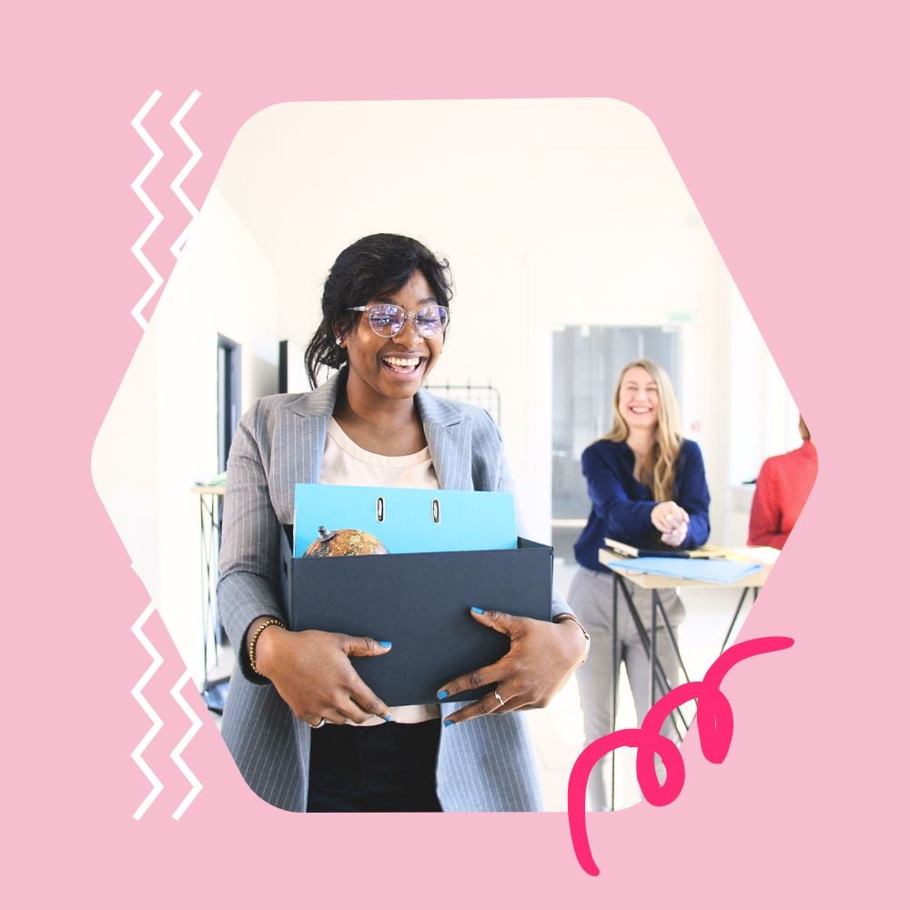 A smiling woman in a gray blazer and glasses holds a box of office supplies, indicating her first day at a new job. In the background, two women, one wearing blue and the other red, smile and cheer her on.