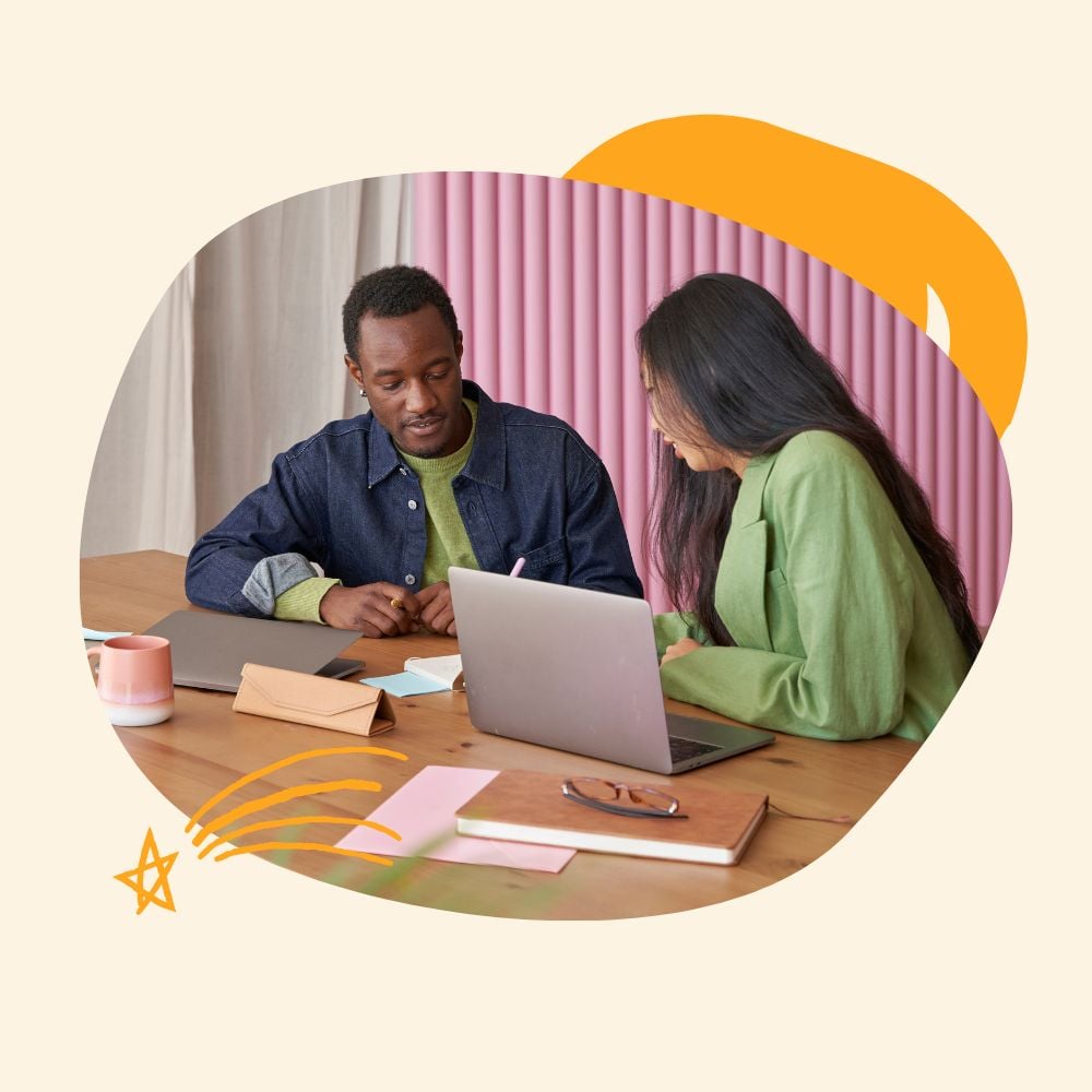 An intern and a manager collaborating at a wooden desk. The intern attentively listens to the manager, who is explaining something on a laptop screen. The background features pink vertical blinds.