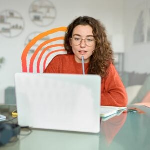 A young woman in glasses and a burnt-orange sweater focused on her laptop, job searching. She holds a pen near her mouth, suggesting deep thought. Her desk, with a notebook and phone, hints at a productive but potentially mistake-prone job search process, like overthinking or missing key details.