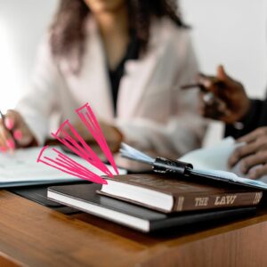 Two people in class at a desk with law-related books, one titled 'The Law,' and papers. One person is holding a pen, and there are pink highlights or marks drawn on the image, emphasizing the focus on law and study materials.